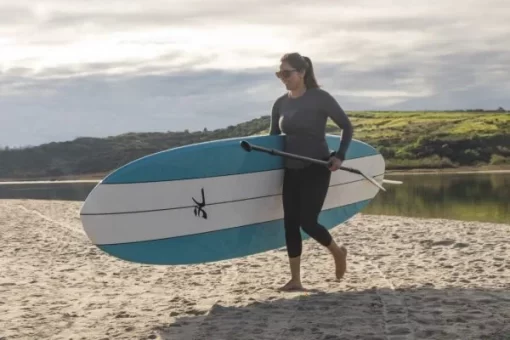A woman carries the Hobie Heritage SUP under 1 arm and the paddle in the other displaying its light weight convenience